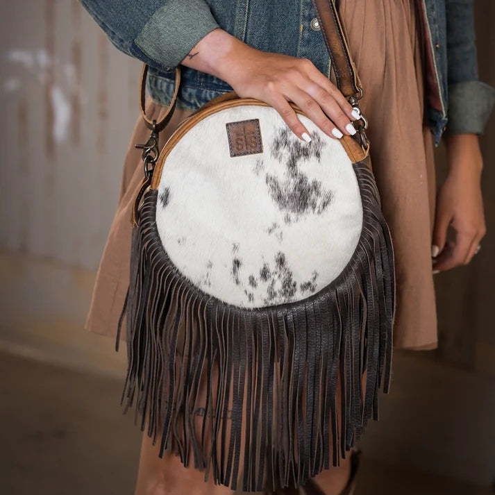 A person wearing a tan vest and denim jacket carries a round fringe crossbody bag with a white faux fur upper section featuring black mottled spots and a dark brown leather fringe.