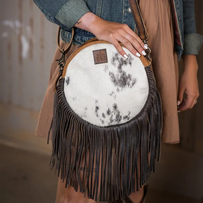 A person wearing a tan vest and denim jacket carries a round fringe crossbody bag with a white faux fur upper section featuring black mottled spots and a dark brown leather fringe.