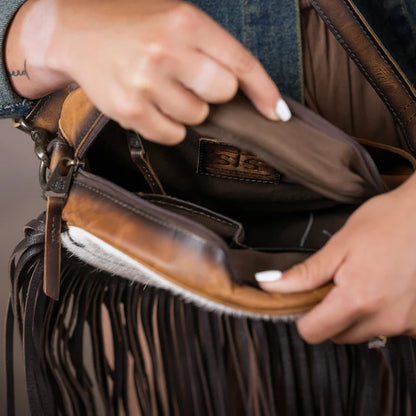 Close-up of a person's hands opening a brown leather fringed shoulder bag, showing the interior lining and a small interior brand patch.