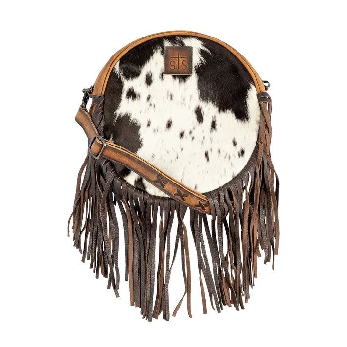 A round cowhide shoulder bag with black and white spotted pattern, brown leather fringe, and a brown leather strap, shown on a plain white background.