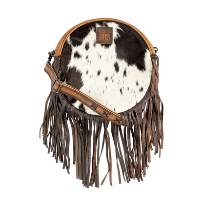 A round cowhide shoulder bag with black and white spotted pattern, brown leather fringe, and a brown leather strap, shown on a plain white background.