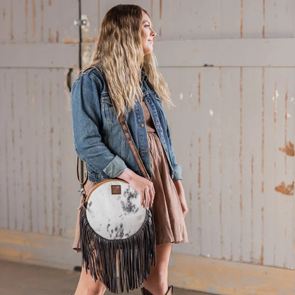 A woman wearing a denim jacket and brown dress carries a round fringe crossbody bag with a white and black marbled cowhide front and dark brown leather fringe, standing in front of a rustic wooden wall.