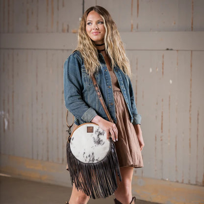 A woman wearing a denim jacket and brown dress carries a round fringe crossbody bag with a white and black marbled cowhide top and black fringe. She is standing in front of a rustic wooden wall.