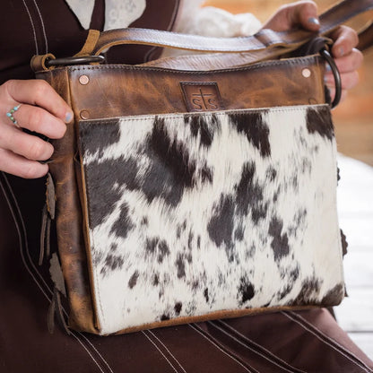 A close-up of a handbag with a brown leather trim and a black-and-white cowhide front panel, held by a person wearing a brown dress.