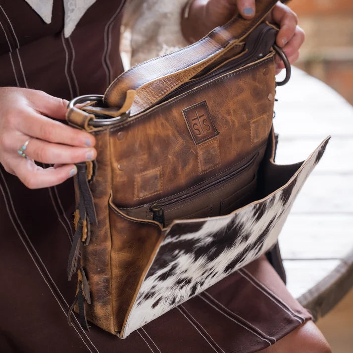 A person holds open a brown leather shoulder bag with a black-and-white cowhide panel on the front, showing the interior compartments. The person is wearing a brown dress, and the setting appears to be a rustic indoor environment.
