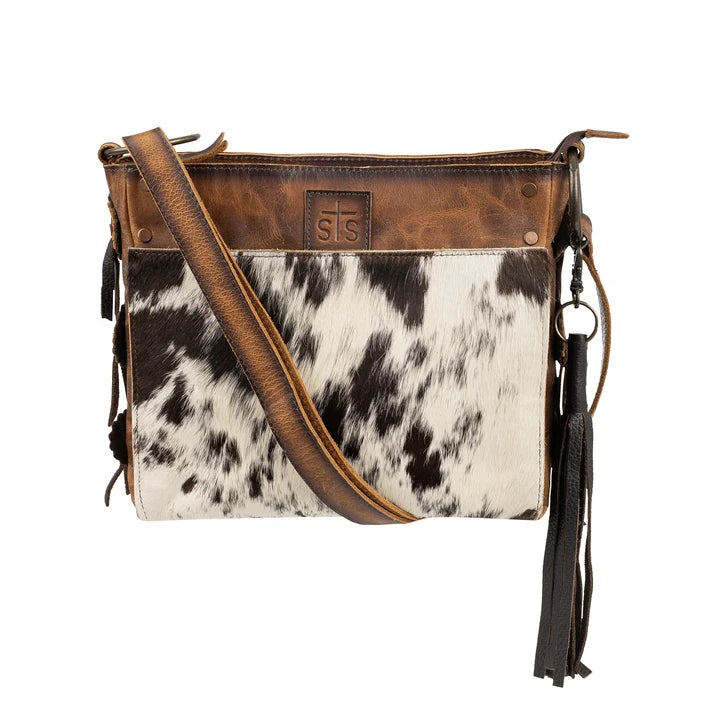 A western-style crossbody bag featuring a brown leather top panel and strap, a black and white cowhide body, and black leather tassels on a white background.