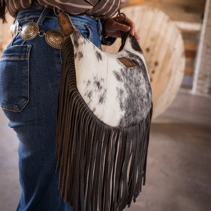 A person wearing blue jeans and a brown striped top carries a slouchy shoulder bag made from cowhide with dark brown fringe. The bag has a white and gray spotted hide top and dark brown leather fringe, photographed in a rustic indoor setting.