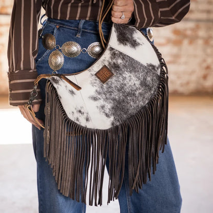 A cowhide and leather fringe shoulder bag worn crossbody over blue jeans, styled with silver concho-style belt hardware and a brown striped shirt. The bag features a white and black cowhide body with dark brown leather fringe and a small brown leather brand patch.