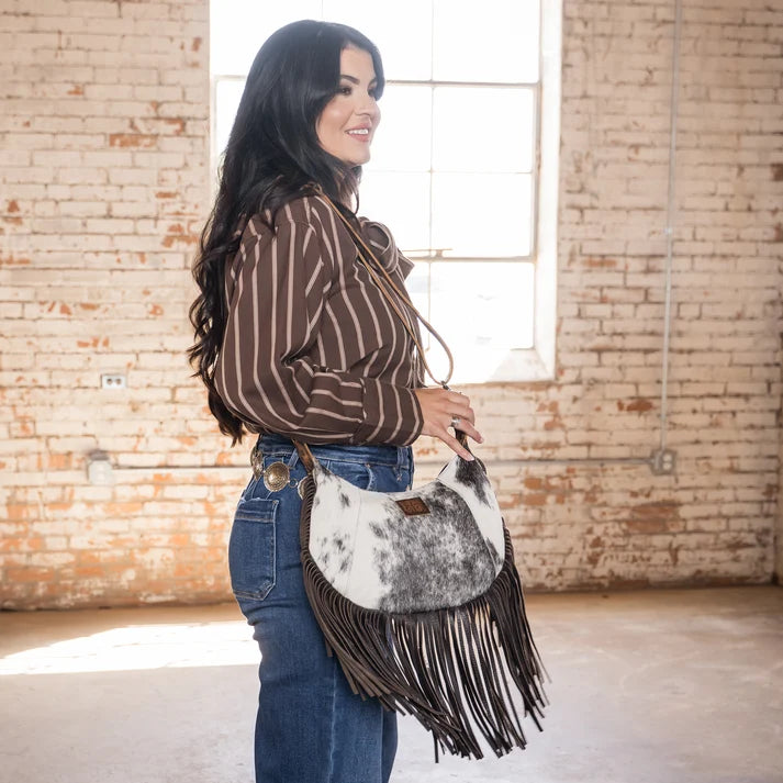 A woman wearing a brown and white striped shirt and blue jeans carries a cowhide fringe shoulder bag in a bright, brick-walled room.