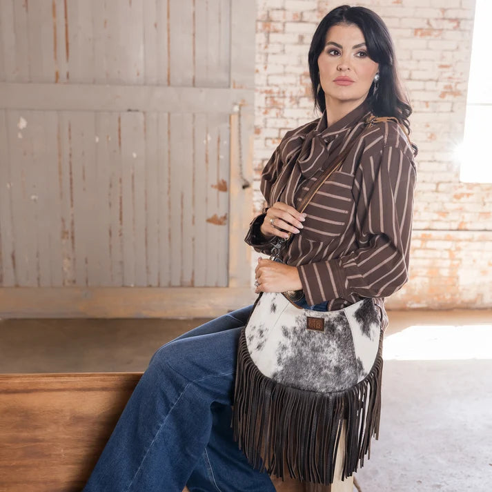 A woman wearing a brown and white striped shirt and blue jeans sits on a wooden bench in a rustic indoor setting, holding a cowhide fringe crossbody bag.