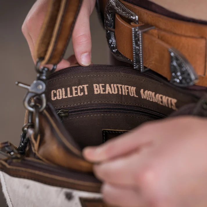 Close-up of a brown leather bag being opened by hands, showing the interior zipper with the embroidered text 'COLLECT BEAUTIFUL MOMENTS' on the inside lining.
