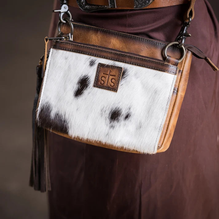 A close-up of a small crossbody bag worn against a brown garment. The bag has a white and black cowhide front panel, tan leather trim, metal rings, and a small brown leather patch with the initials 'sts'.