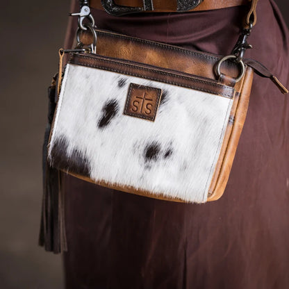 A close-up of a small crossbody bag worn against a brown garment. The bag has a white and black cowhide front panel, tan leather trim, metal rings, and a small brown leather patch with the initials 'sts'.