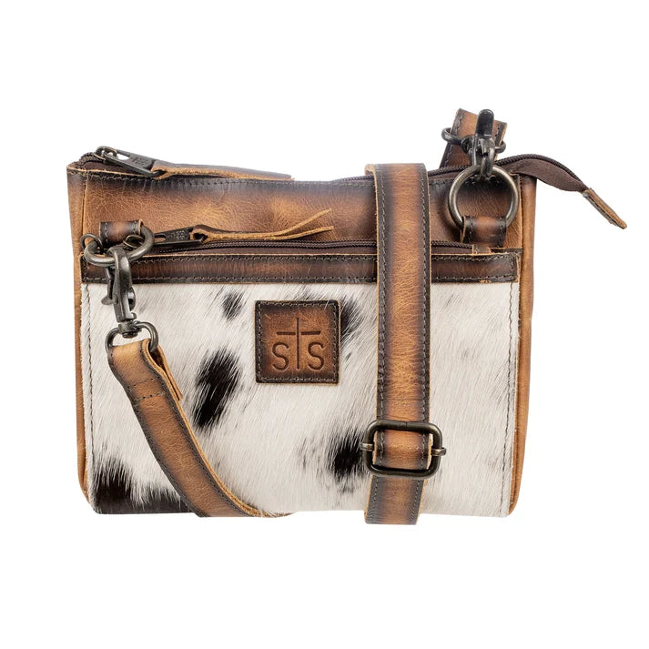 A small crossbody bag with a brown leather top and straps, a white cowhide lower panel with black spots, and a brown leather patch with the initials 'STS' on the front, shown on a white background.