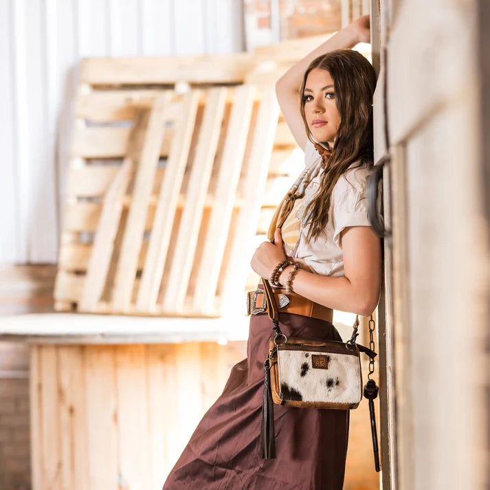 A woman wearing a white t-shirt and brown skirt leans against a wooden wall, carrying a brown and white cow-print shoulder bag with a tassel.