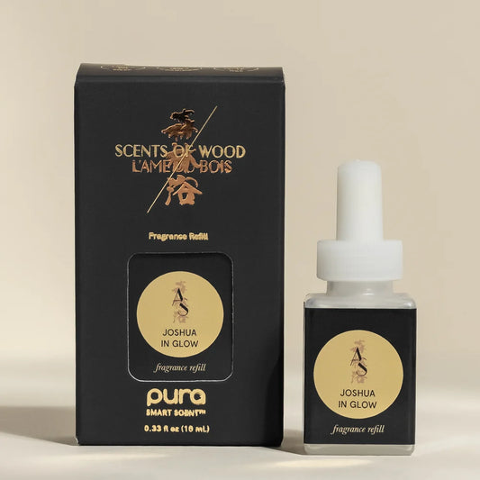 A bottle of Pura Smart Scent 'Joshua In Glow' fragrance refill next to its black packaging box with gold lettering, set against a solid beige background.