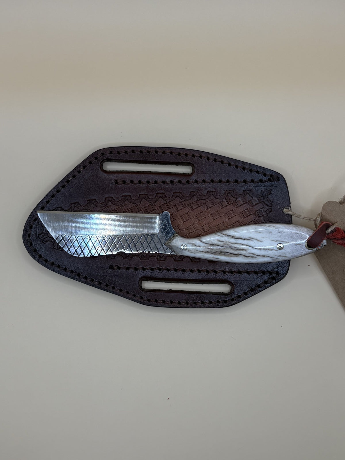 A fixed-blade knife with a textured silver blade and brown handle, partially inserted into a dark brown leather sheath on a beige background.
