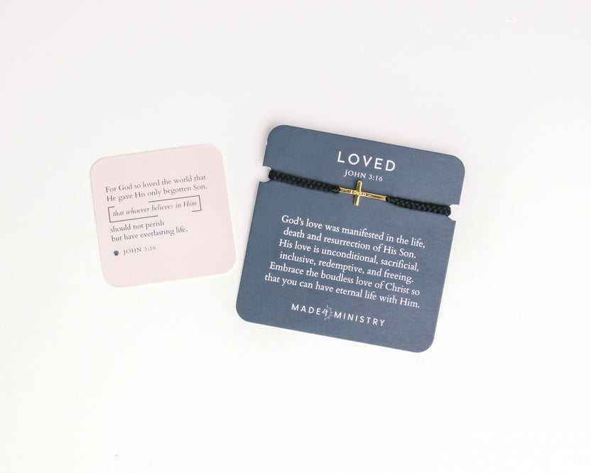 Bracelet with cross design and 'LOVED' text on a gray card, with pink card in the background.