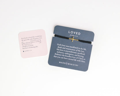Bracelet with cross design and 'LOVED' text on a gray card, with pink card in the background.