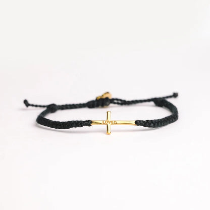 A black braided cord bracelet with a small gold cross charm engraved with the word 'LOVE', displayed on a clean white background.