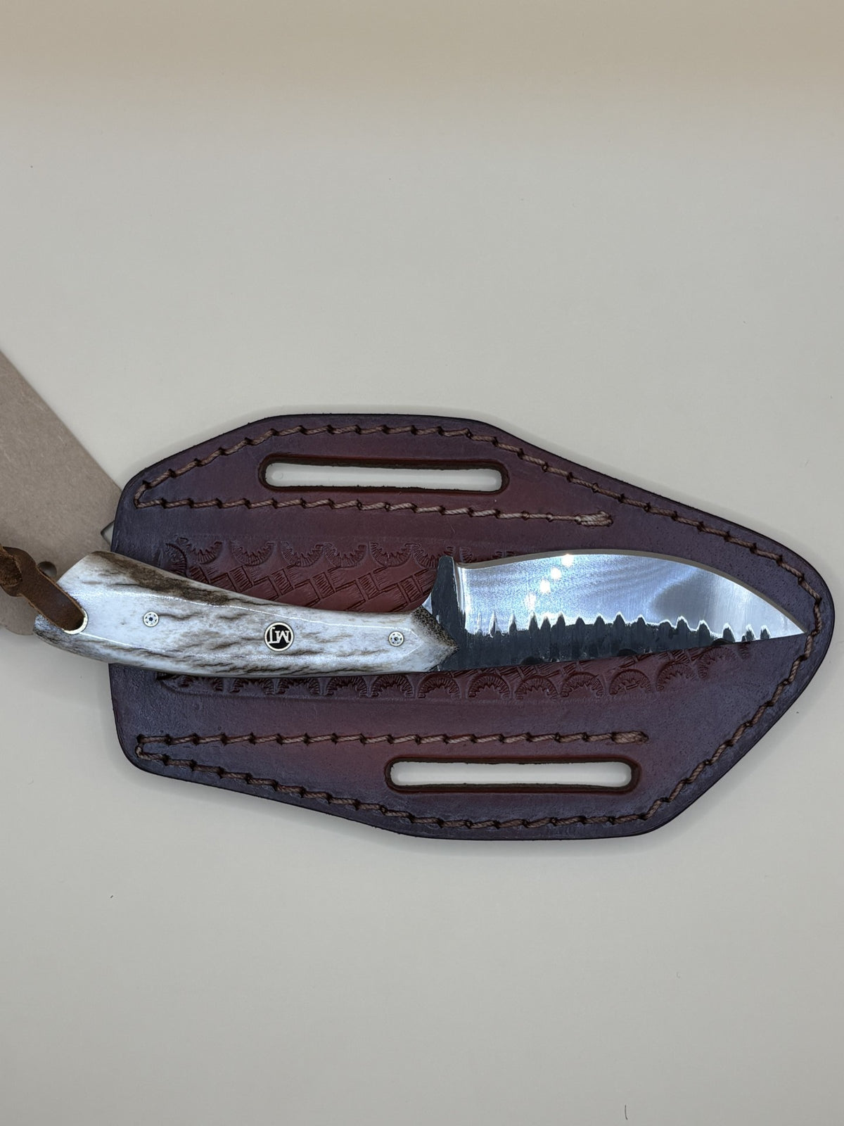 A fixed-blade knife with a textured silver handle and a serrated steel blade, partially tucked into a dark brown leather sheath with cutout slots, shown on a plain light beige background.