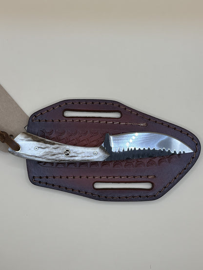 A fixed-blade knife with a textured silver handle and a serrated steel blade, partially tucked into a dark brown leather sheath with cutout slots, shown on a plain light beige background.