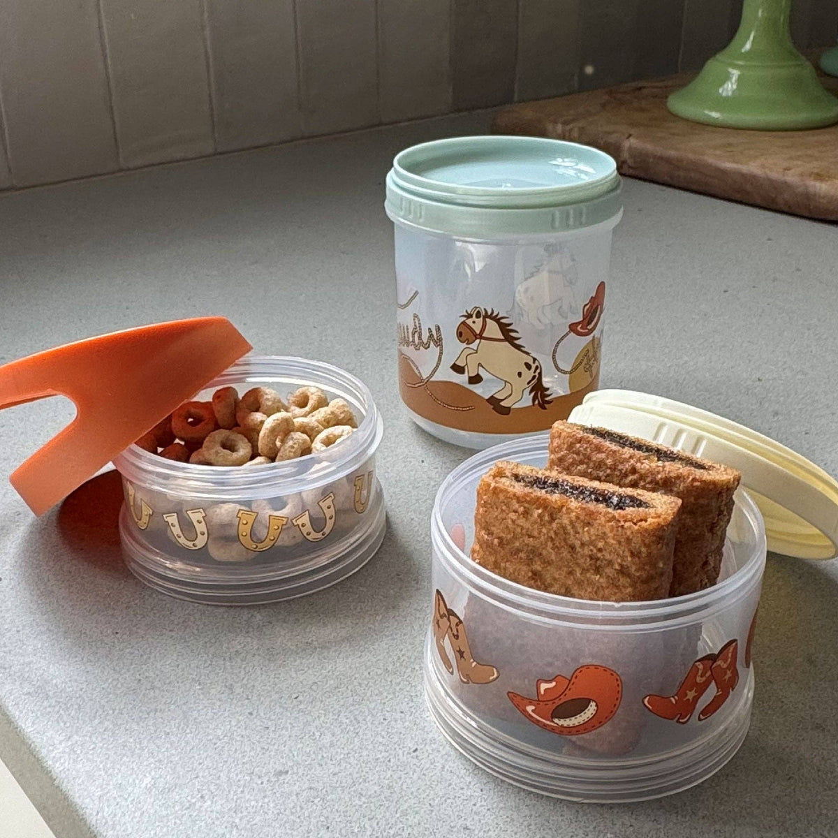 A set of three stacked snack containers with a boots and Howdy motif, in orange, green, and clear colors, with animal illustrations.