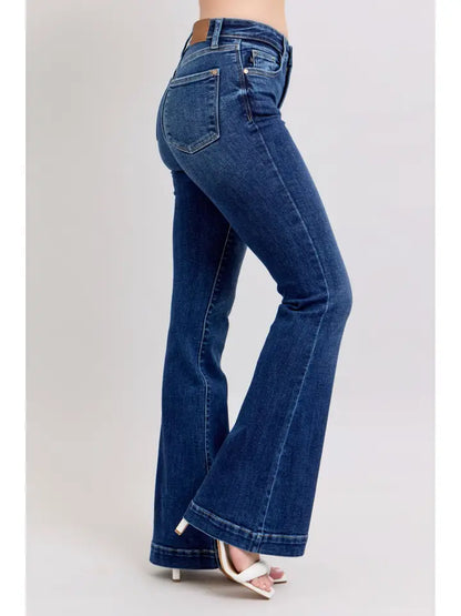 A person wearing dark blue flare jeans with a brown leather waistband detail and white heeled sandals, shown from the waist down against a light gray studio background.
